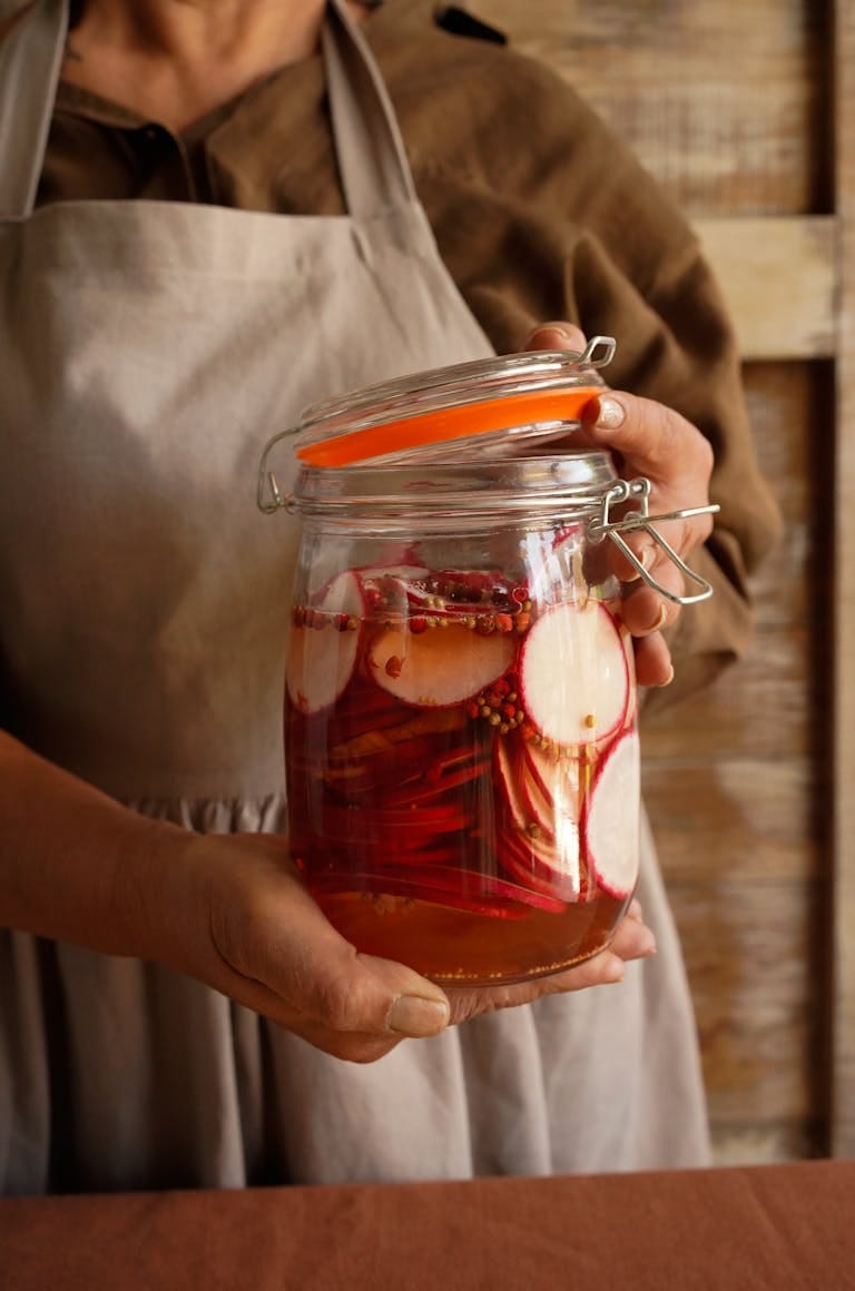 A woman in an apron holds a jar of homemade pickled vegetables.