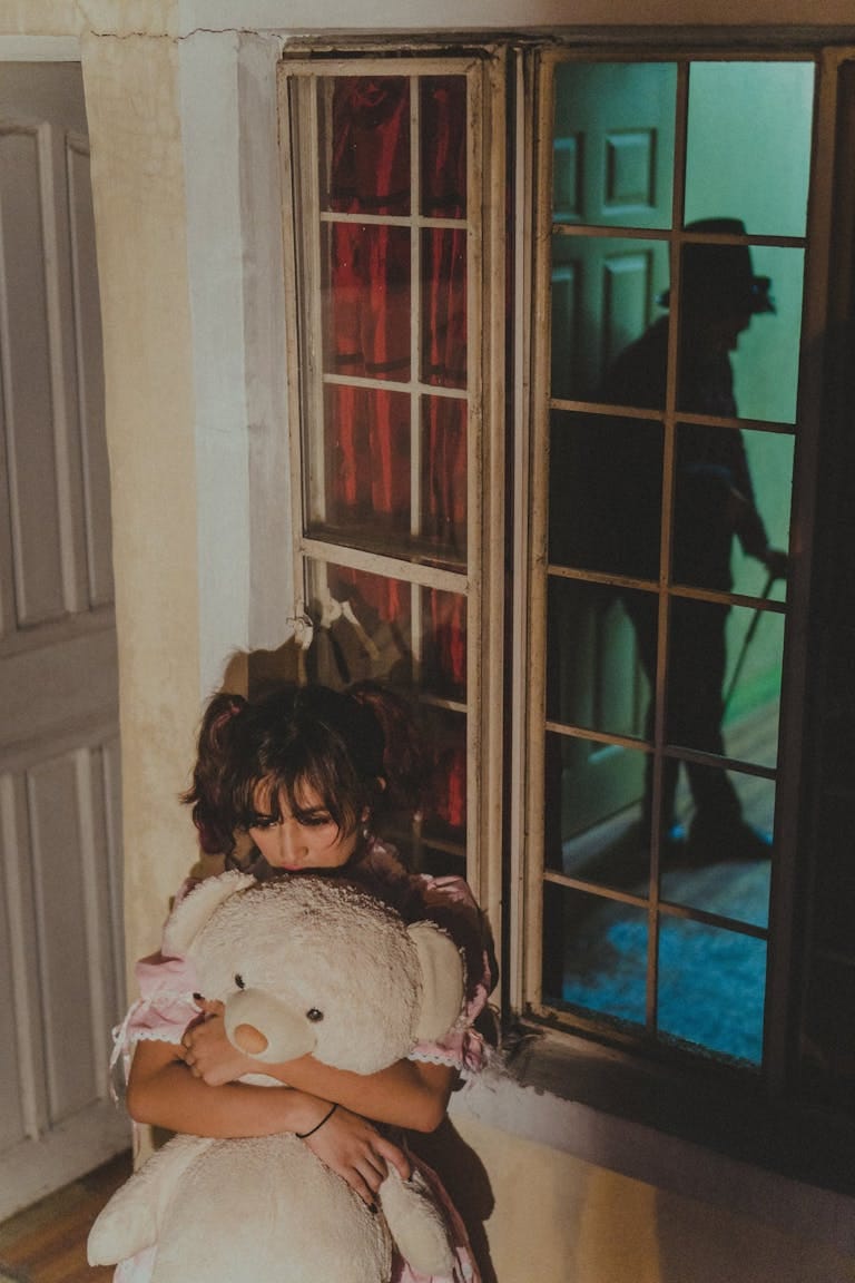 A child in distress hugging a teddy bear with a menacing shadow visible through window.